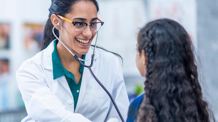 A doctor with long dark hair in a ponytail, glasses, and a white lab coat smiles and checks a child’s vital signs using a stethoscope.